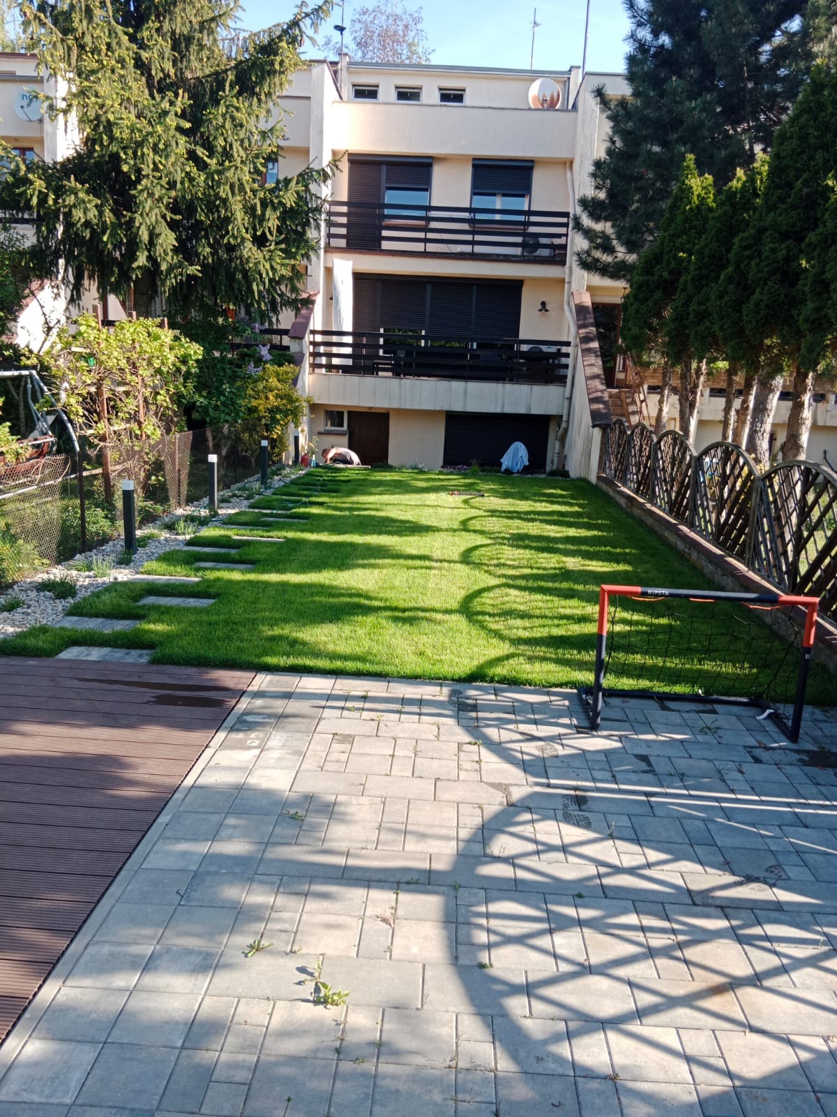 Zadbany trawnik przed domem z kostką brukową i drewnianym tarasem. Widoczne kamienne płyty w trawie, mała bramka do piłki nożnej i ogrodzenie.