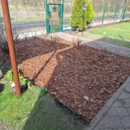 Kwadratowa rabata wysypana korą, obrzeża z tworzywa, trawnik, żółte kwiaty w doniczce przy brązowym słupku, zielona furtka w tle.