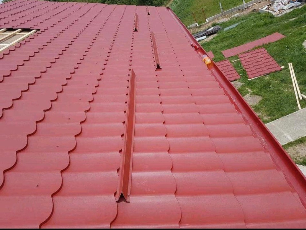 Czerwony dach z blachy trapezowej o falistym wzorze, widoczne elementy rynny i fragment konstrukcji drewnianej w tle.