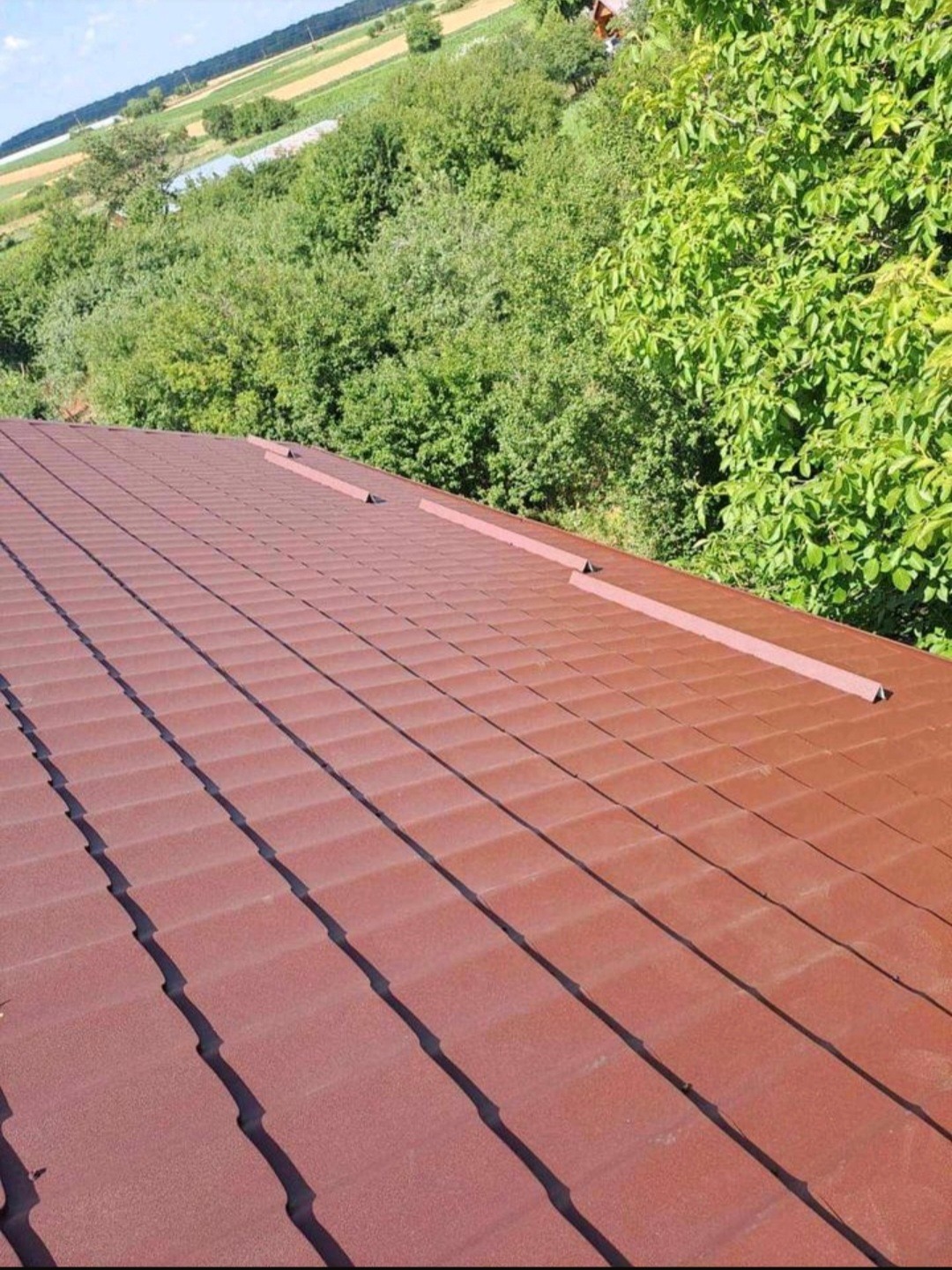 Czerwona blachodachówka na nowym dachu, widok z góry na tle zieleni drzew i pól uprawnych.