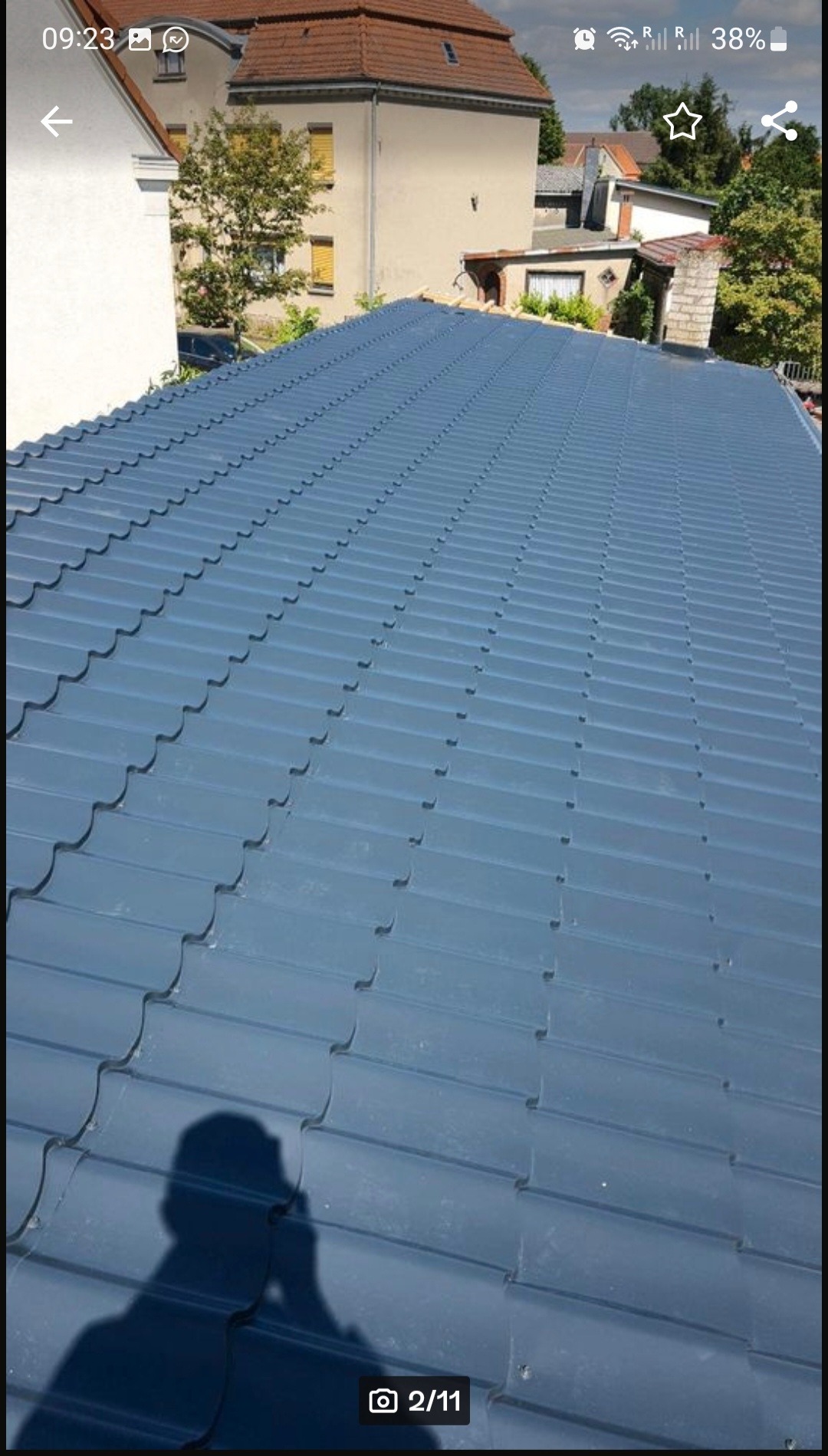 Błękitny dach z blachodachówki, widok z góry, cień osoby robiącej zdjęcie w lewym dolnym rogu, w tle budynki mieszkalne i drzewa.
