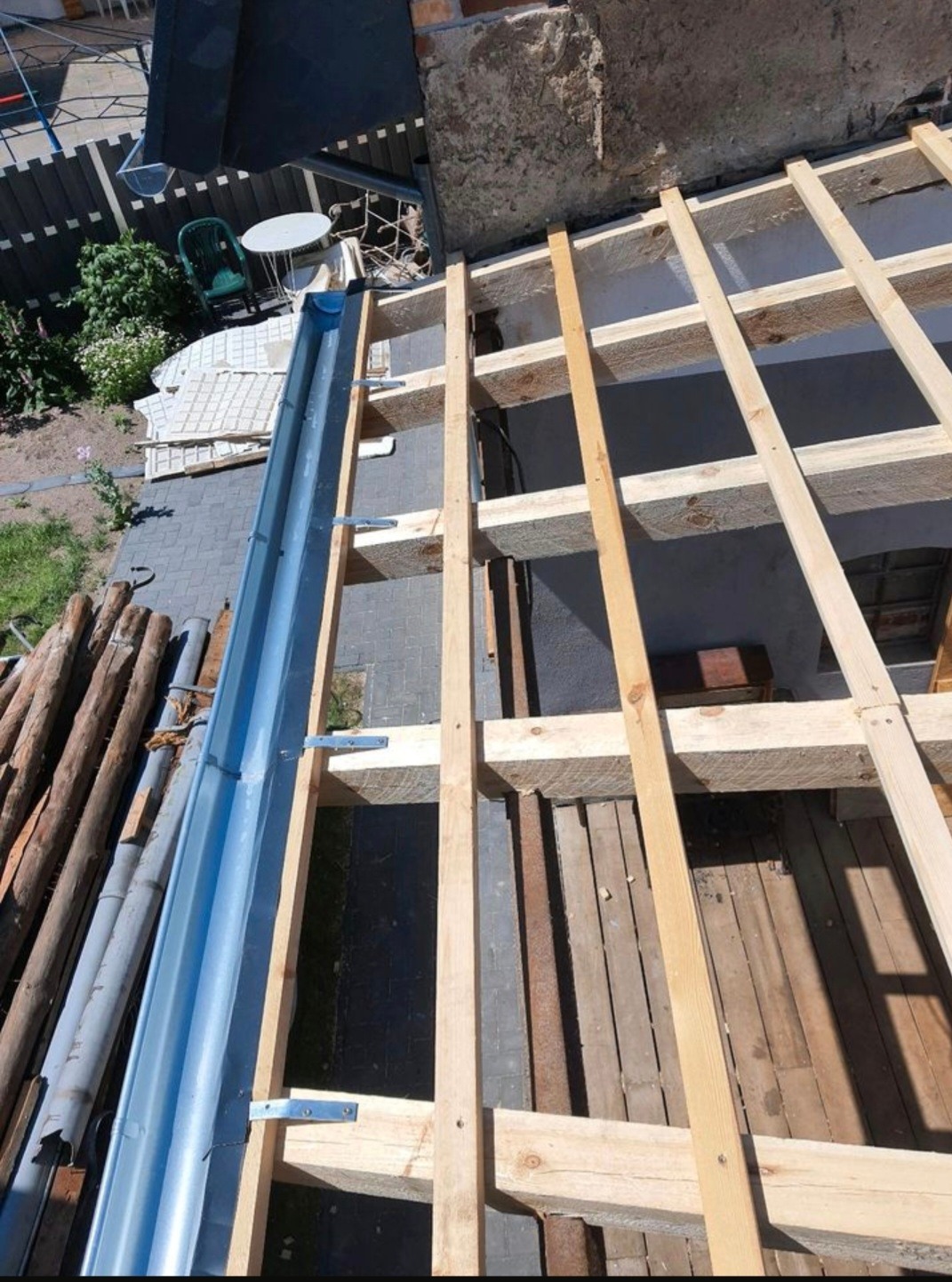 Widok z góry na konstrukcję dachu z drewnianych belek i zamontowaną, błyszczącą, metalową rynną, z widocznymi elementami mocującymi. W tle fragment ogrodu z meblami.