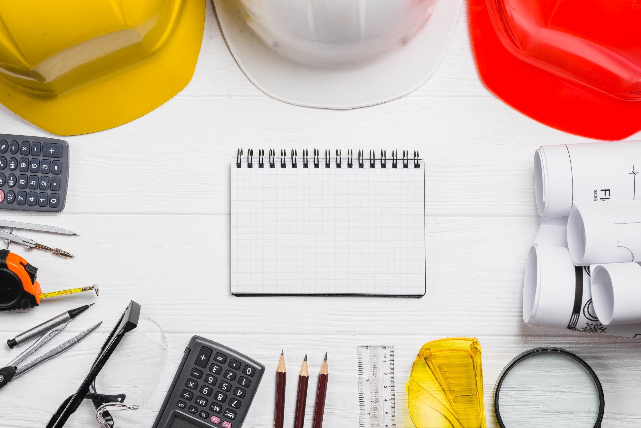 Planowanie robót ziemnych: kask żółty, biały i czerwony, notatnik, kalkulator, miara, ołówki, lupa i zwinięte plany na białym drewnianym stole.