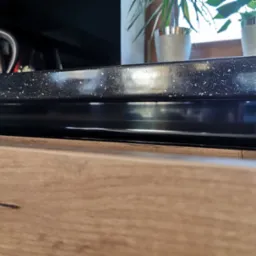 Fragment blatu kuchennego z czarnego, błyszczącego konglomeratu z białymi drobinkami, widoczny front szuflady z drewnopodobnego materiału, w tle czajnik elektryczny i rośliny doniczkowe...