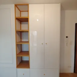 Biała, lakierowana szafa z drewnianym regałem po lewej stronie, wyposażona w złote gałki, stojąca na drewnianej podłodze w jasnym wnętrzu.