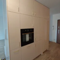 Beżowa zabudowa kuchenna na wymiar z piekarnikiem i złotymi uchwytami, widok z perspektywy podłogi z paneli.