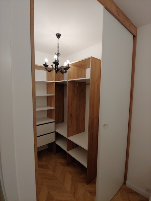Wnętrze szafy wnękowej z przesuwnymi, białymi drzwiami, drewnianymi półkami i szufladami, oświetlone dekoracyjnym, czarnym żyrandolem, widok na parkiet ułożony w jodełkę.