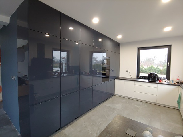 Nowoczesna kuchnia z ciemnoszarymi, lakierowanymi szafkami sięgającymi sufitu, białymi szafkami dolnymi i czarnym blatem, widok na okno z zielenią na zewnątrz.