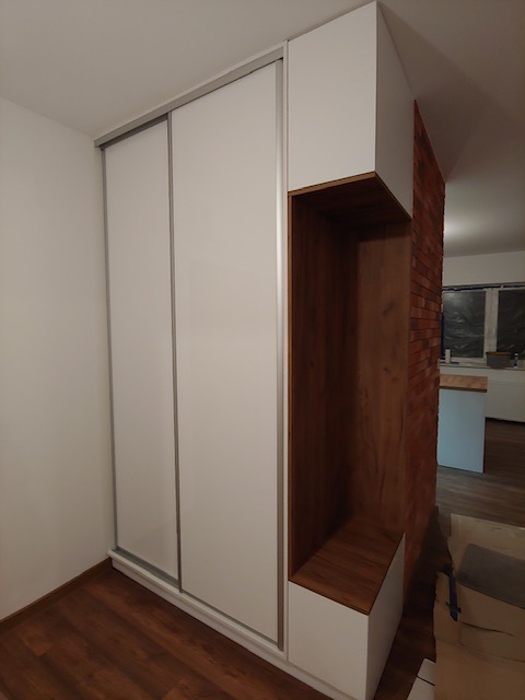 Biała szafa przesuwna z drewnianym regałem wbudowanym w ścianę z cegły, widoczne drewniane panele podłogowe.