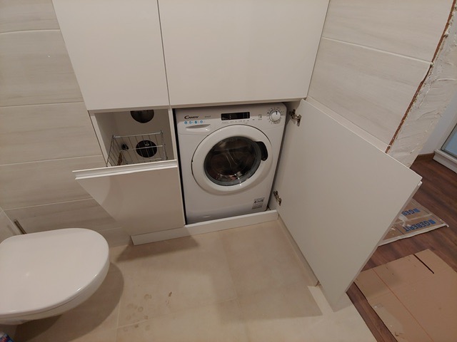 Biała pralka Candy w zabudowie meblowej w łazience, z otwartymi drzwiczkami szafki po prawej stronie i szufladą z koszem na pranie po lewej, obok toaleta.