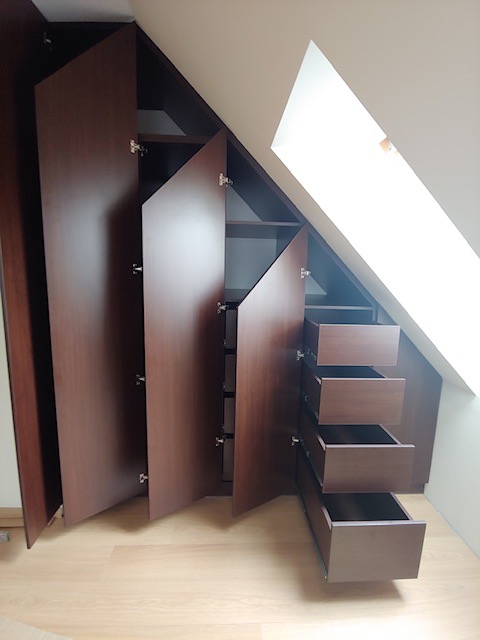 Brązowa zabudowa szafy wnękowej na poddaszu ze skośnym sufitem, z otwartymi drzwiami i wysuniętymi szufladami, ukazująca półki i wnętrze.