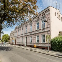 Elewacja kamienicy w Wrześni, z widocznymi detalami architektonicznymi, takimi jak ozdobne okna i gzymsy, sfotografowana z poziomu ulicy w słoneczny dzień.