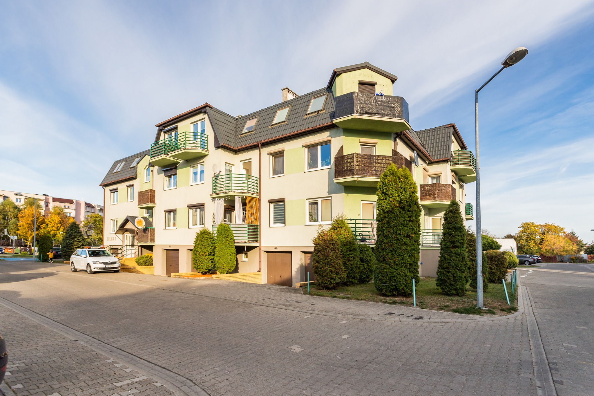 Wielopiętrowy budynek mieszkalny z balkonami, szarą dachówką i zielonymi akcentami kolorystycznymi, otoczony zielenią i ulicą z kostki brukowej, z zaparkowanym białym samochodem osobowym.