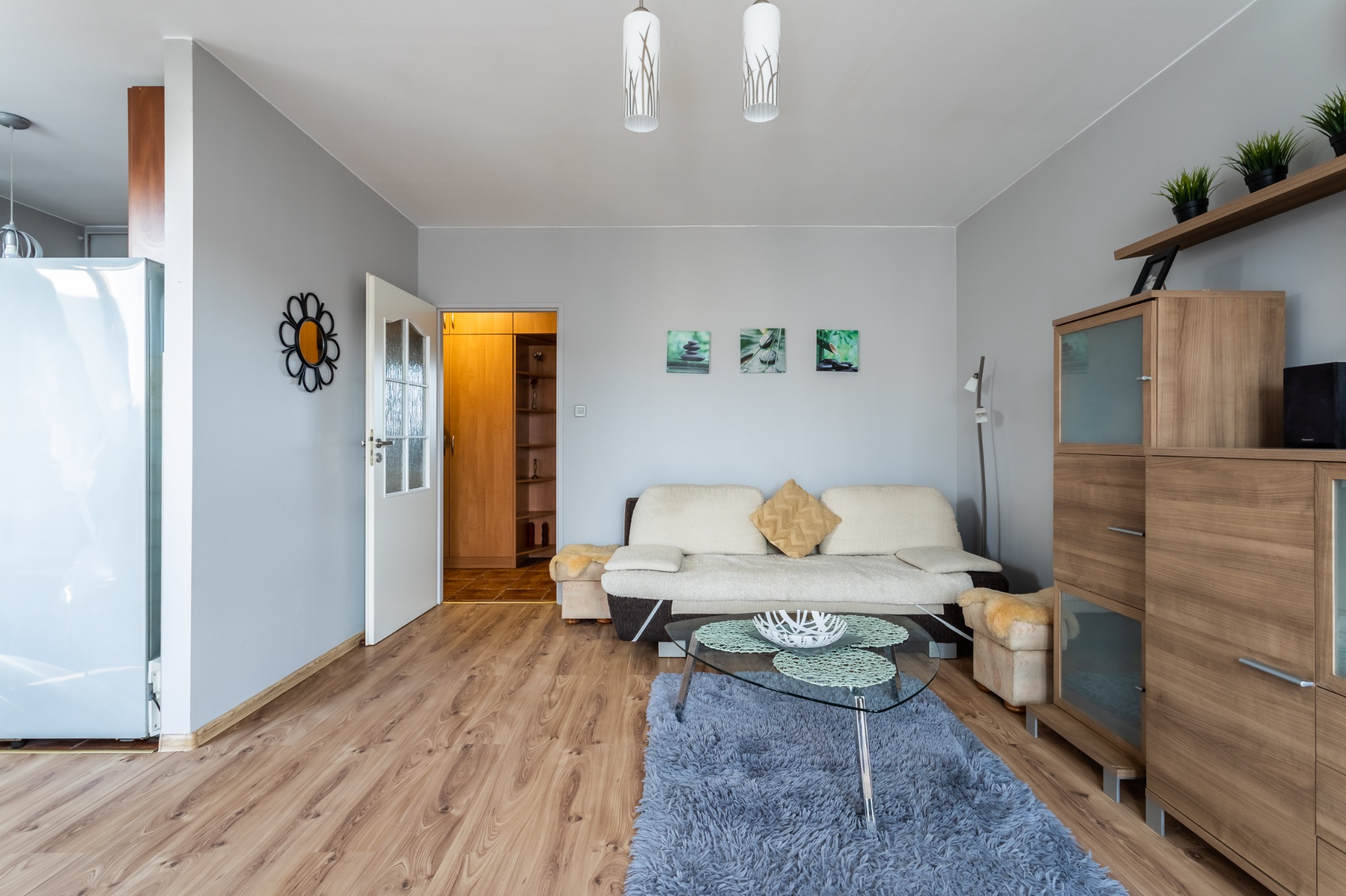 Jasne wnętrze salonu z drewnianą podłogą, szarą ścianą, beżową kanapą i szarym dywanem, z dekoracjami w postaci obrazków i kwiatów w doniczkach.