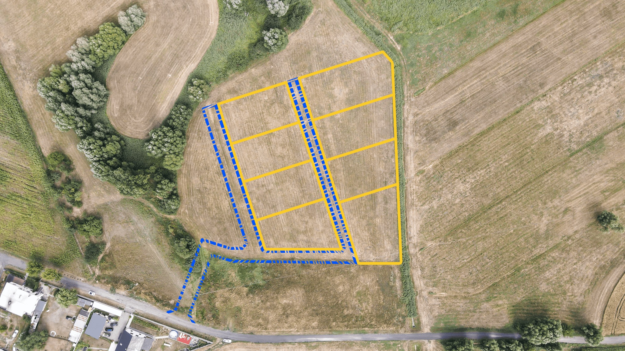 Widok z lotu ptaka na działkę budowlaną z naniesionymi liniami podziału w kolorze żółtym i liniami przyłączy w kolorze niebieskim, przylegającą do zabudowań mieszkalnych i drogi.