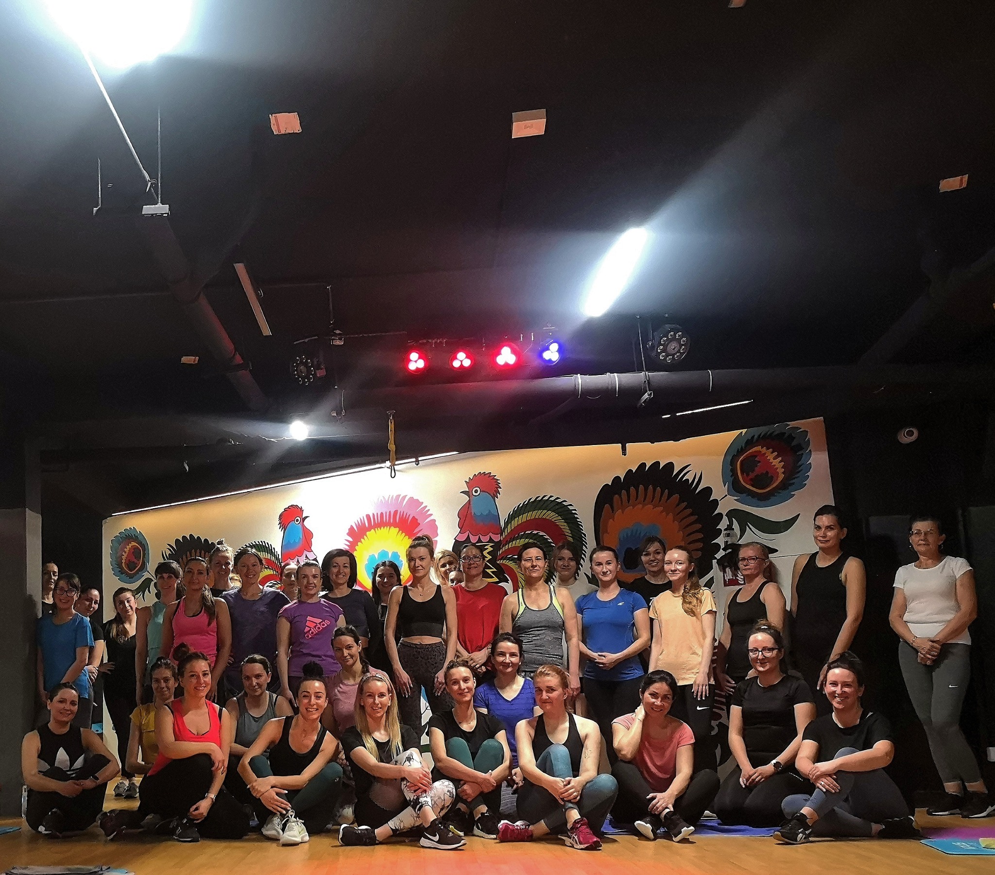 Duża grupa kobiet w strojach sportowych pozuje do zdjęcia w sali z ludowym motywem koguta na ścianie, oświetlonej reflektorami.
