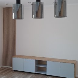 JS.MEBLE Janusz Słomski - Nowoczesne wnętrze z drewnianą ścianą, szafką RTV z szarymi frontami i drewnianym blatem, oraz trzema wiszącymi lampami z czarnymi elementami i szklanymi kloszami.