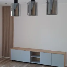 Nowoczesne wnętrze z drewnianą ścianą, szafką RTV z szarymi frontami i drewnianym blatem, oraz trzema wiszącymi lampami z czarnymi elementami i szklanymi kloszami.