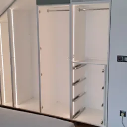 Biała szafa wnękowa z oświetleniem LED w sypialni na poddaszu, widoczne szyny do szuflad i narzędzia na podłodze.