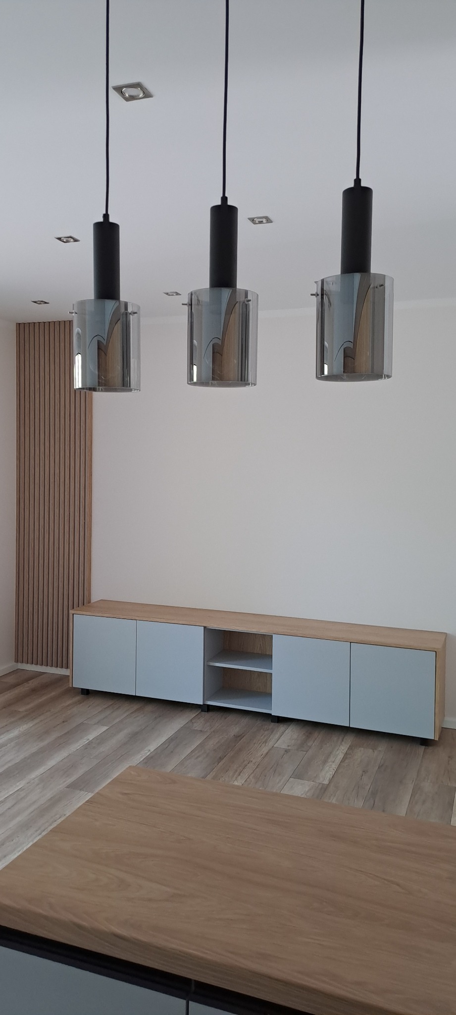 Nowoczesne wnętrze z drewnianą ścianą, szafką RTV z szarymi frontami i drewnianym blatem, oraz trzema wiszącymi lampami z czarnymi elementami i szklanymi kloszami.