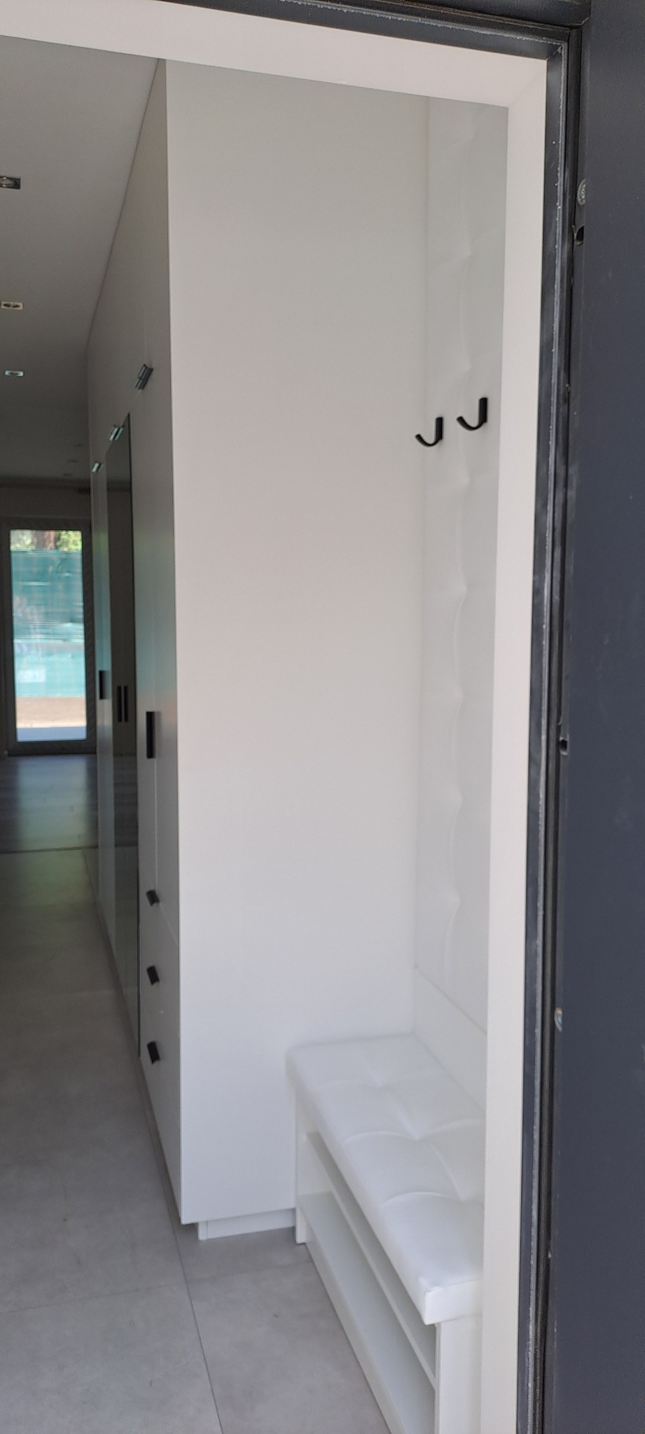 Wnętrze przedpokoju z białą szafą wnękową z siedziskiem i wieszakami, widok z korytarza w stronę otwartych drzwi wejściowych.