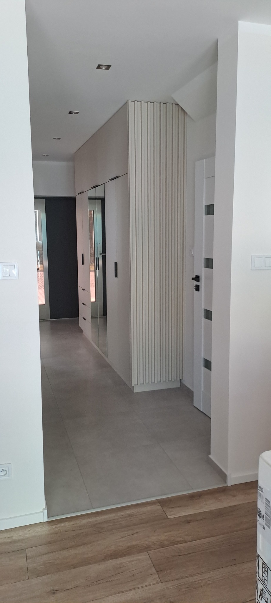 Nowoczesny przedpokój z zabudowaną szafą z lustrami i ryflowanym panelem dekoracyjnym, szara podłoga z płytek i drewnopodobne panele, białe ściany i drzwi.