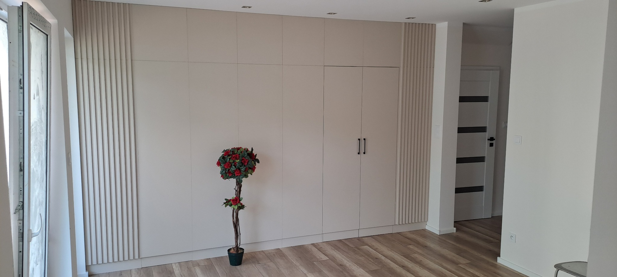 Duża, jasna szafa wnękowa z dekoracyjnymi panelami pionowymi, uzupełniona sztucznym drzewkiem w doniczce na tle parkietu.