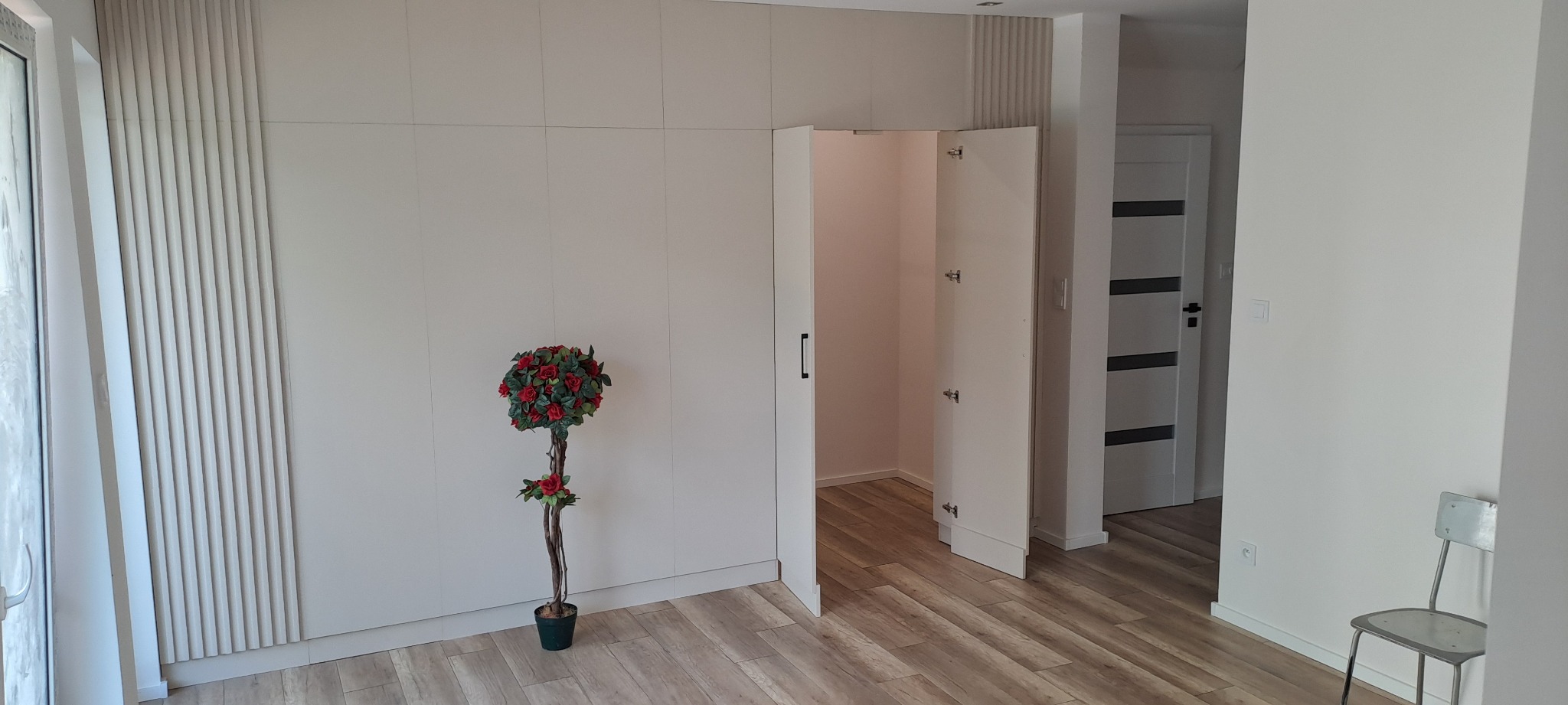 Wnętrze z dużą, białą szafą wnękową z otwartymi drzwiami, dekoracyjnym panelem ściennym i sztucznym drzewkiem w doniczce.