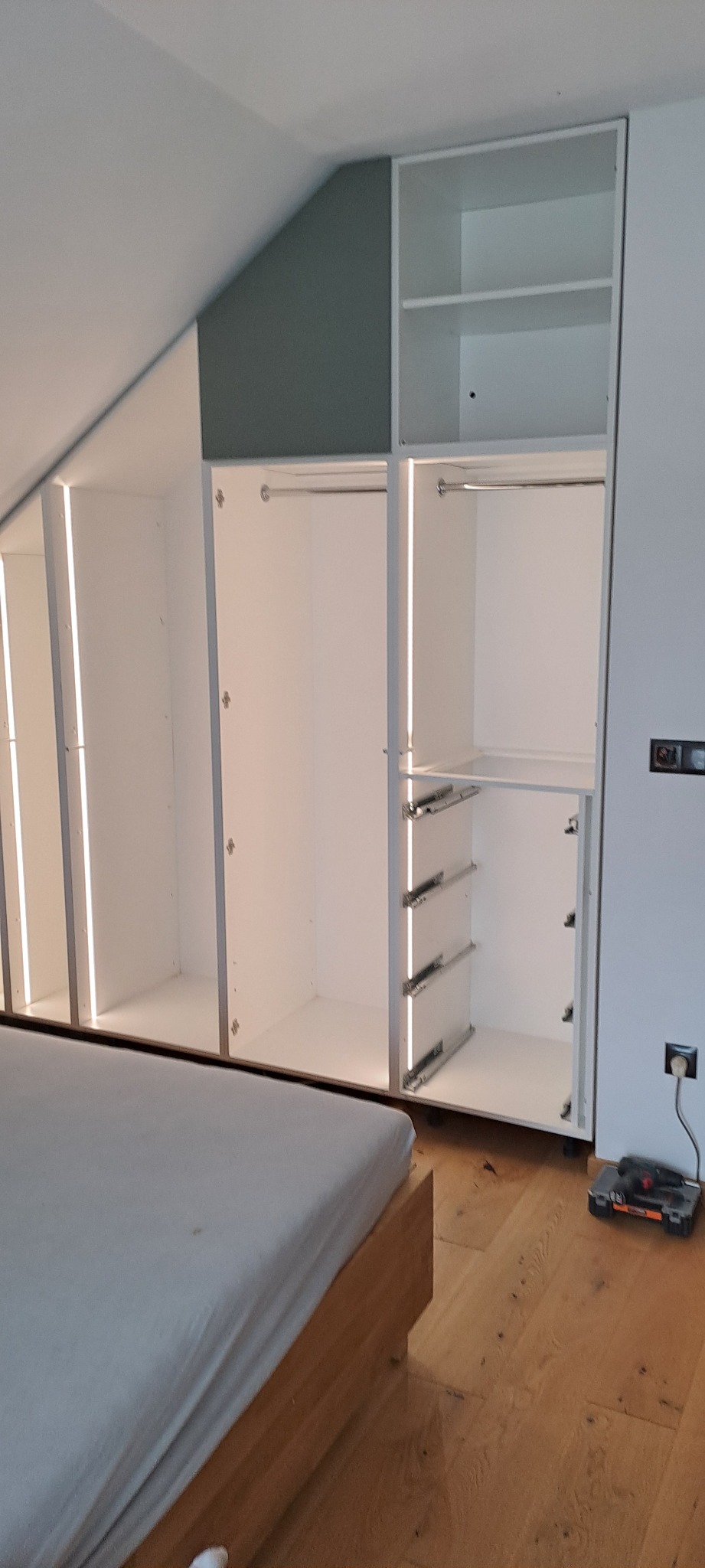 Biała szafa wnękowa z oświetleniem LED w sypialni na poddaszu, widoczne szyny do szuflad i narzędzia na podłodze.