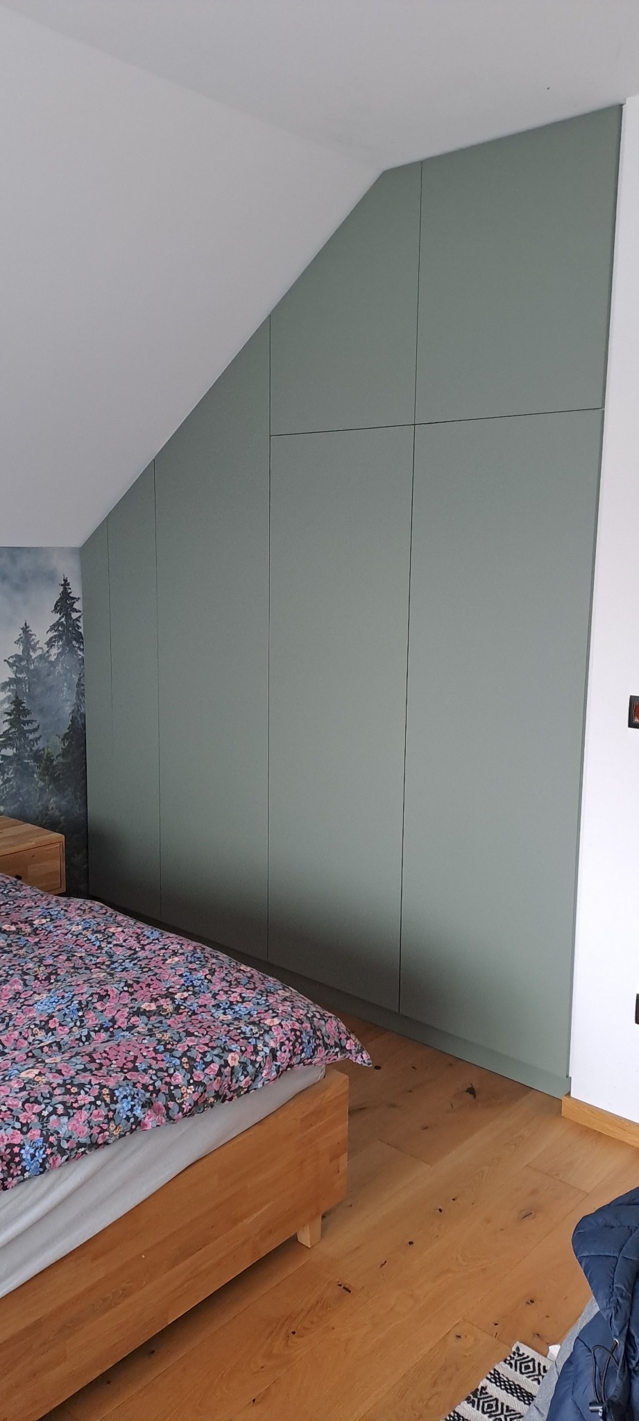 Zielona szafa wnękowa z frontami bez uchwytów, dopasowana do skosu poddasza, widoczna z boku łóżka z kwiecistą pościelą, na drewnianej podłodze.