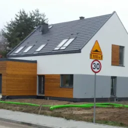 Nowoczesny dom z drewnianą elewacją, szarą dachówką i oknami dachowymi. Widoczny znak drogowy i zielone rury na trawniku. Częściowo wykończony teren.