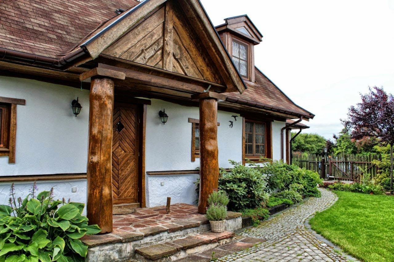 Biały dom z drewnianym gankiem, zdobionymi kolumnami i brązowym dachem. Widoczny kamienny chodnik i zadbany ogród. Styl rustykalny.