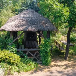 Zadaszona altana z grillem i stołem w otoczeniu zieleni, widok z zewnątrz na murowany grill.