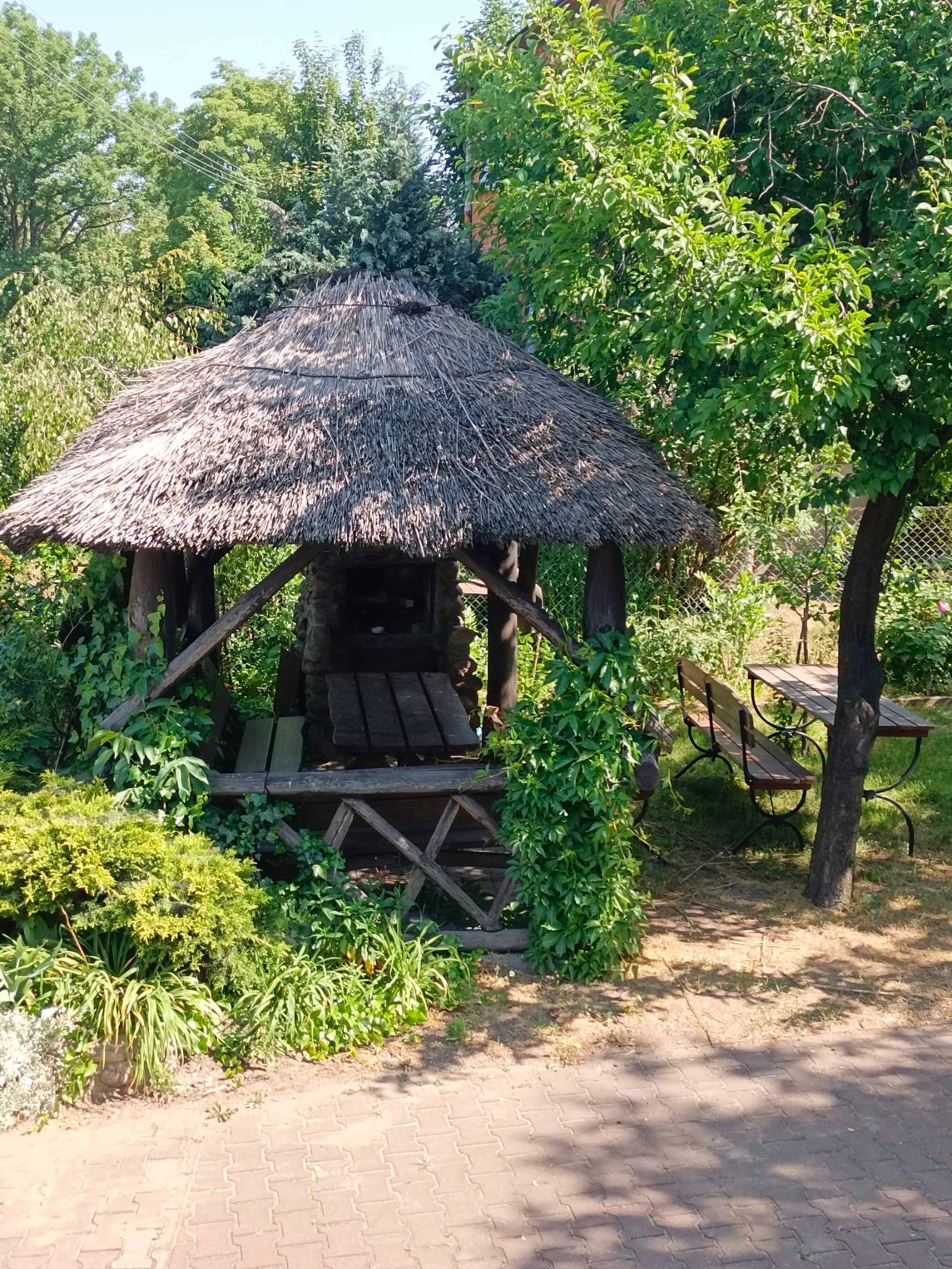 Zadaszona altana z grillem i stołem w otoczeniu zieleni, widok z zewnątrz na murowany grill.