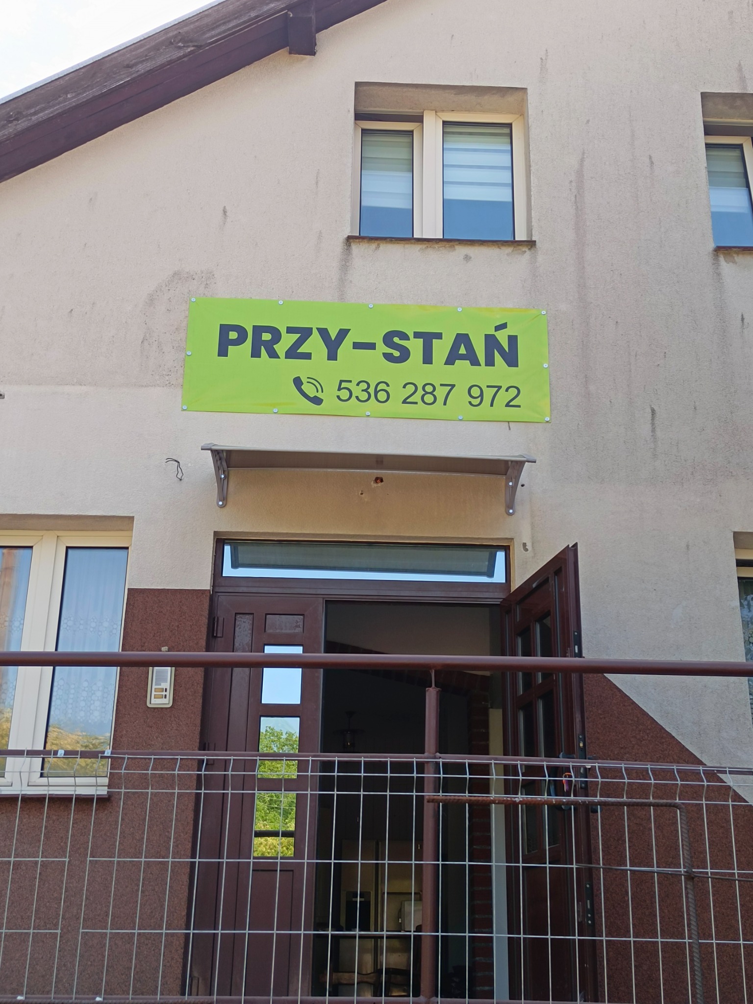 Zewnętrzny widok budynku z zielonym szyldem 'PRZY-STAŃ' i numerem telefonu nad wejściem, widziany zza metalowego ogrodzenia balkonu.