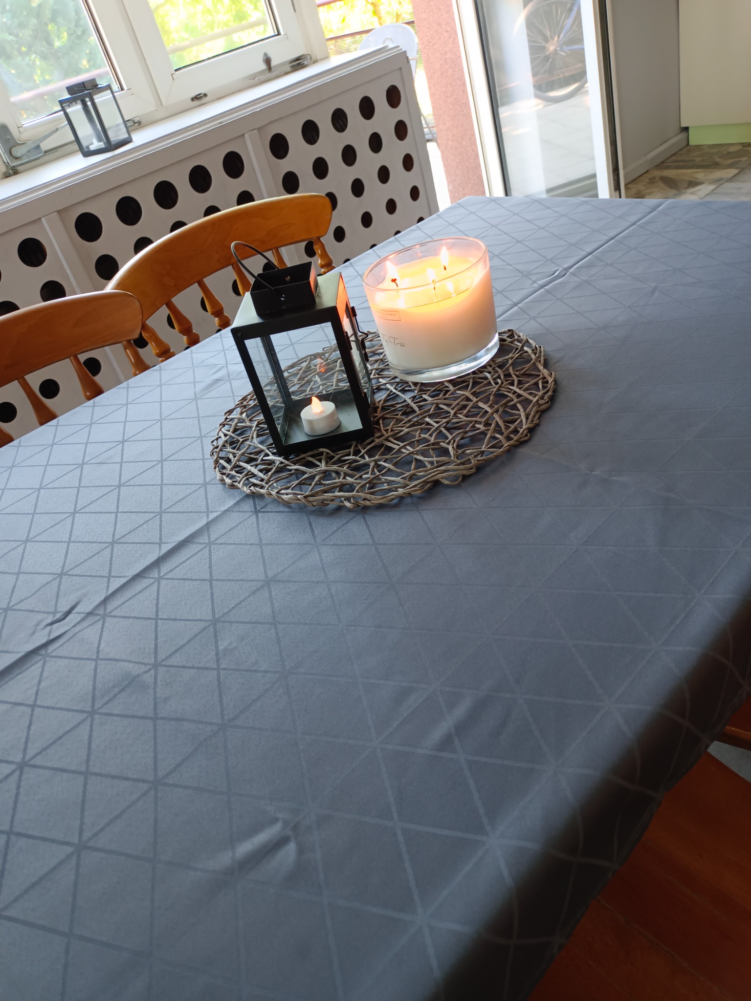 Stół przykryty szarym obrusem w geometryczny wzór, na nim lampion ze świecą oraz duża świeca w szklanym pojemniku, ustawione na podkładce z wikliny.