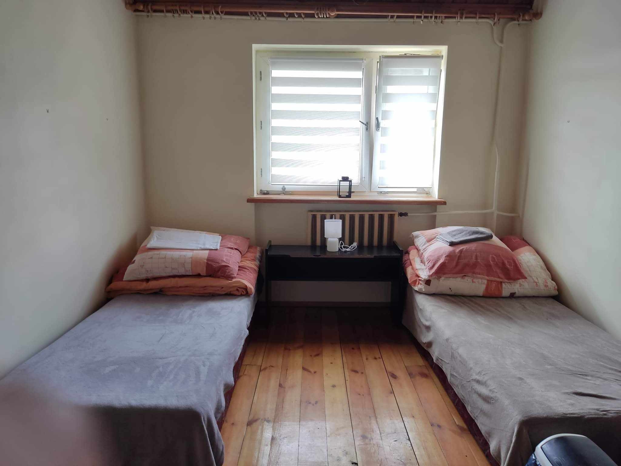 Pokój z dwoma łóżkami w Giewartowie, drewniana podłoga, okno z żaluzjami, stolik nocny z lampką.