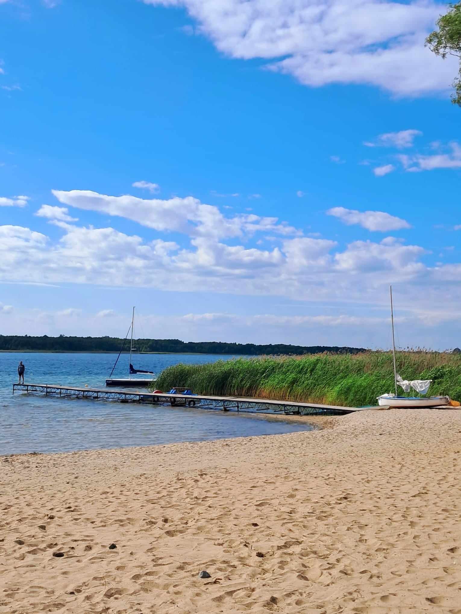 Piaszczysta plaża nad jeziorem z drewnianym pomostem, na którym stoi mężczyzna, w tle trzciny, zacumowana łódka i błękitne niebo z chmurami.
