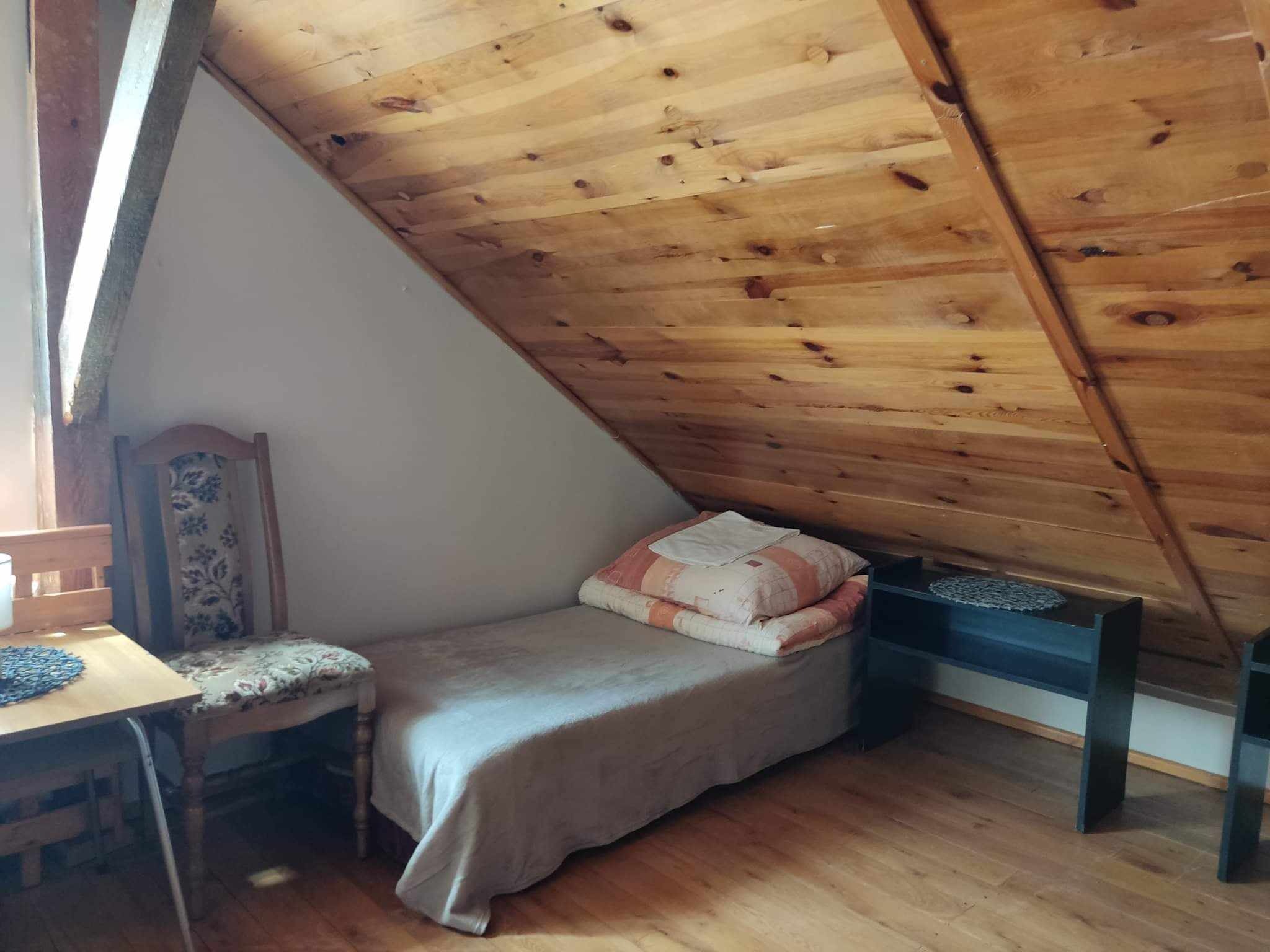 Pokój na poddaszu z drewnianym wykończeniem sufitu i podłogi, pojedyncze łóżko z pościelą w odcieniach beżu i pomarańczy, krzesło z tapicerką w kwiaty przy drewnianym stoliku, mała czarna półka.