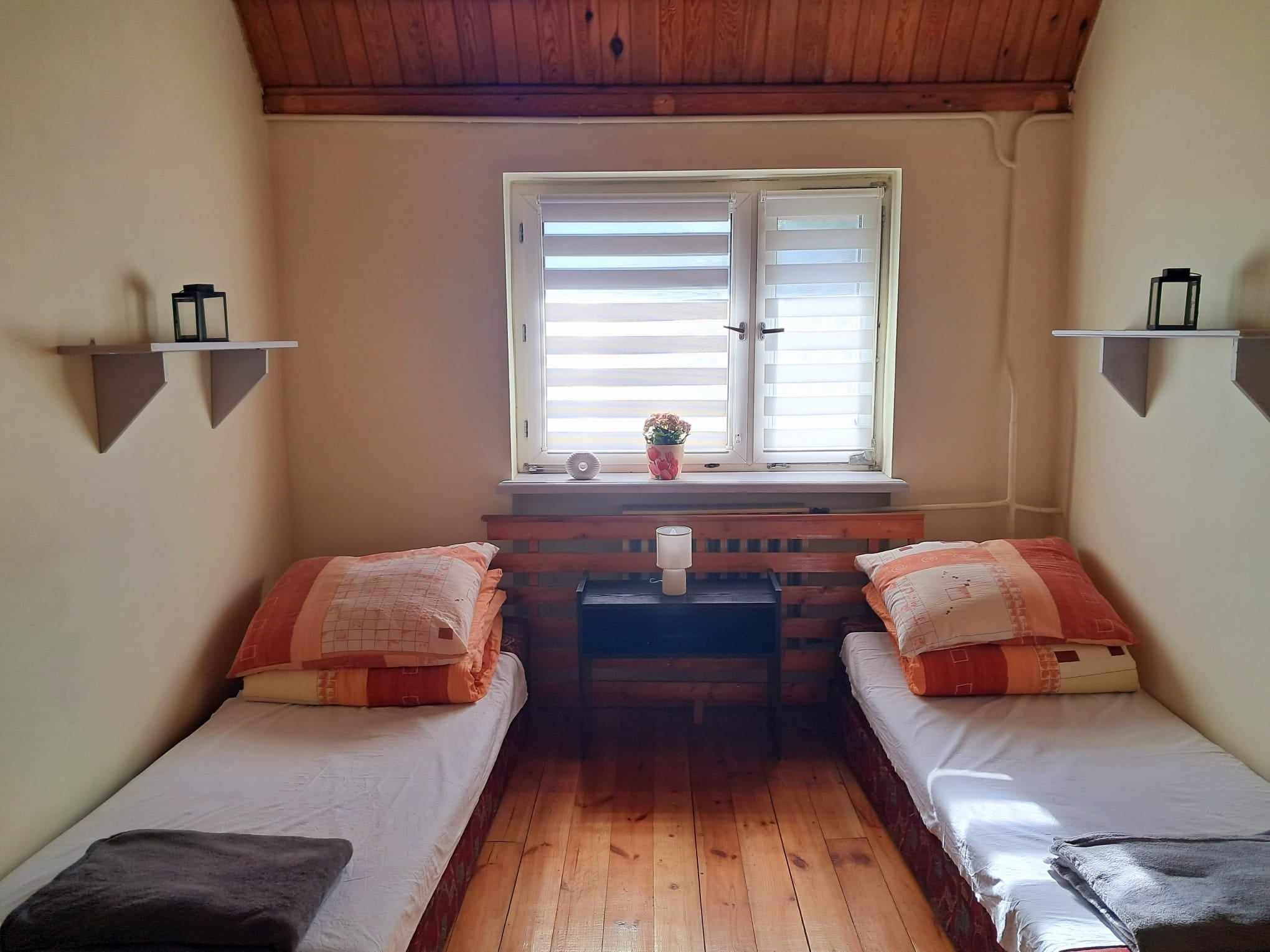 Pokój z dwoma pojedynczymi łóżkami, drewnianą podłogą i sufitem, małym stolikiem nocnym między łóżkami i oknem z żaluzjami.