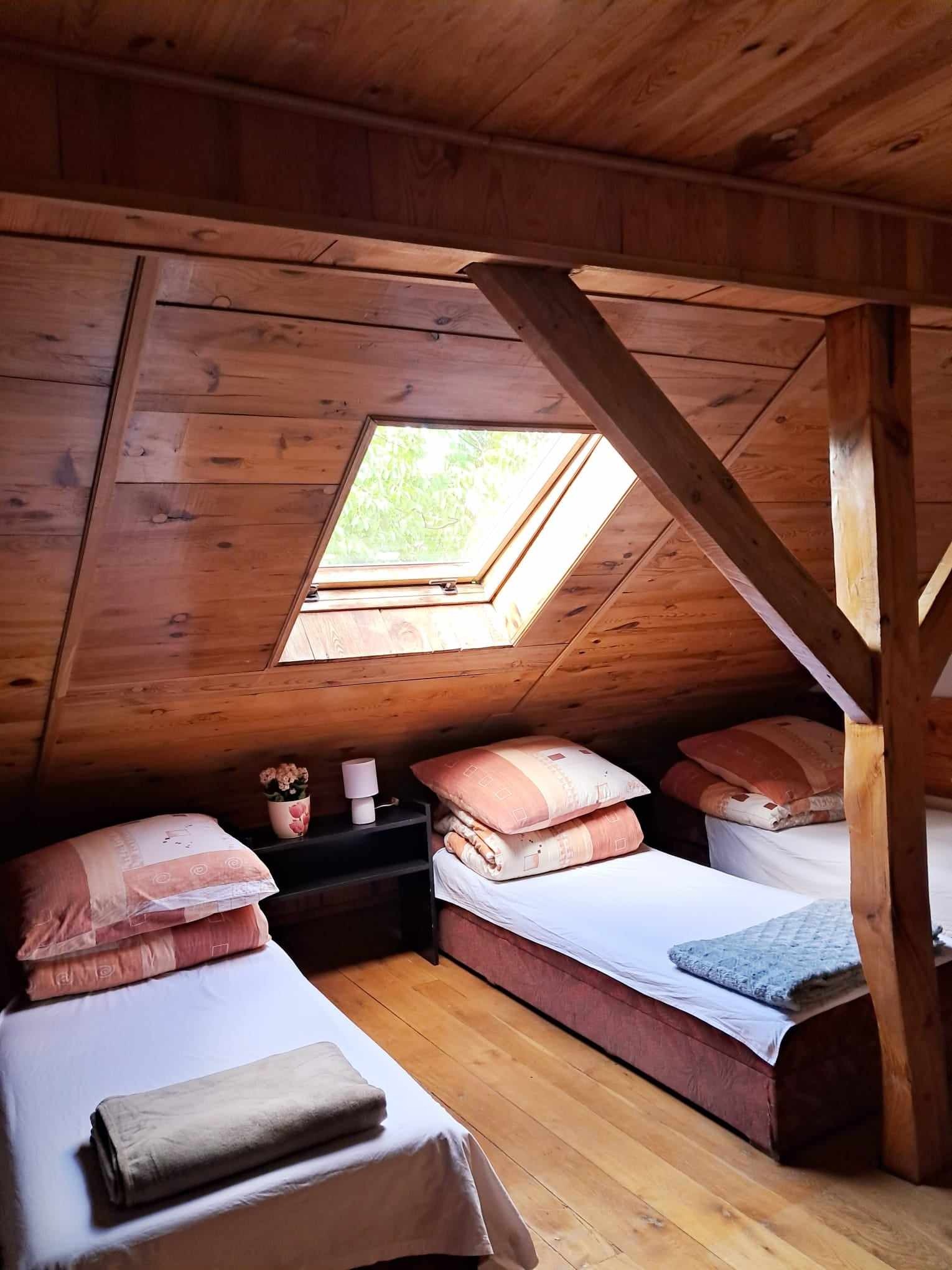 Drewniany pokój na poddaszu z trzema pojedynczymi łóżkami, oknem dachowym i widocznymi belkami konstrukcyjnymi.