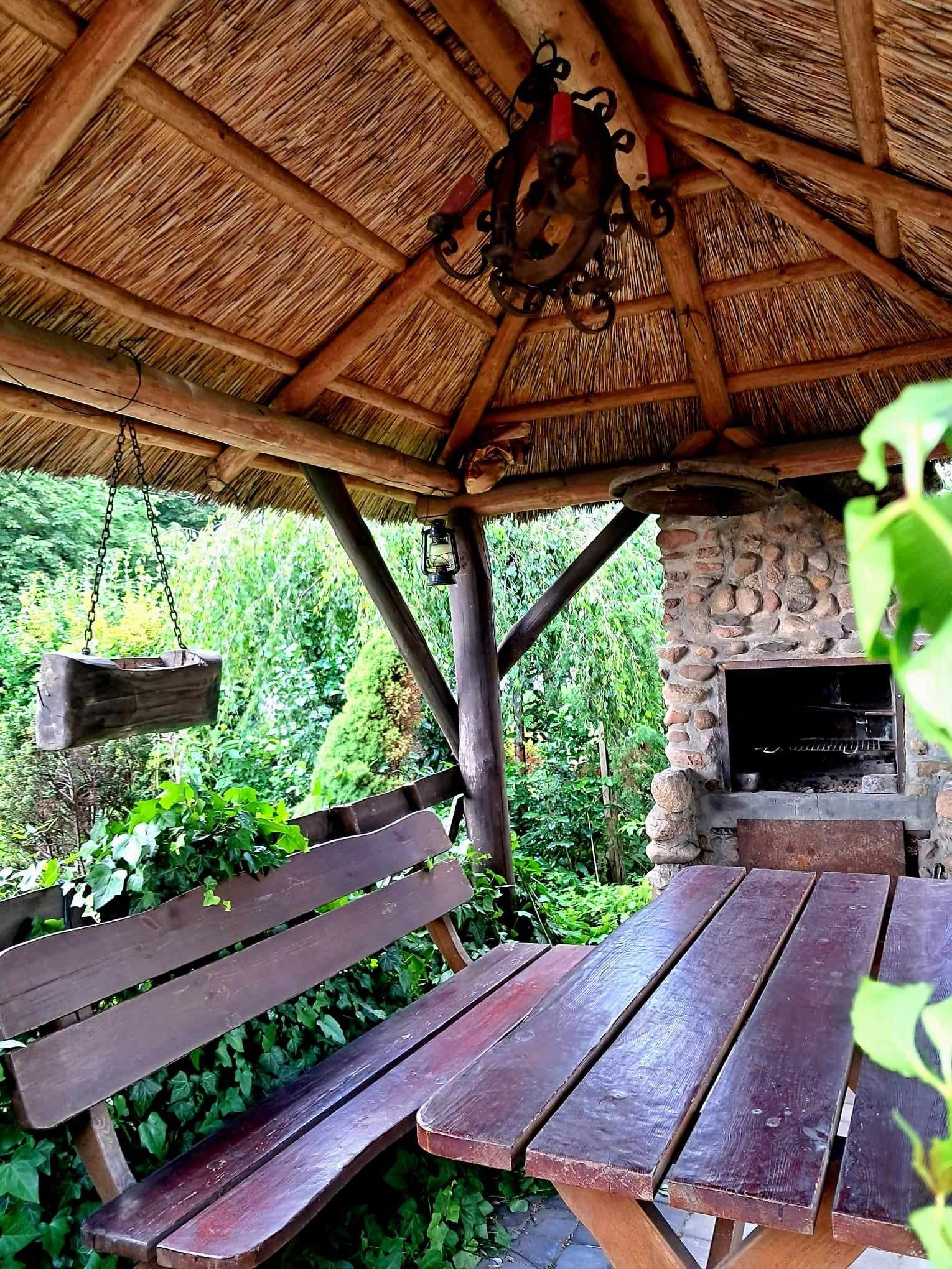 Drewniana altana z krytym strzechą dachem, kamiennym grillem, drewnianym stołem i ławką, otoczona zielenią ogrodu.