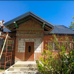 Paweł Statuch BuildKraft - Pracownik na drabinie montuje elementy dachu na nowym domu z czerwonej cegły w słoneczny dzień.