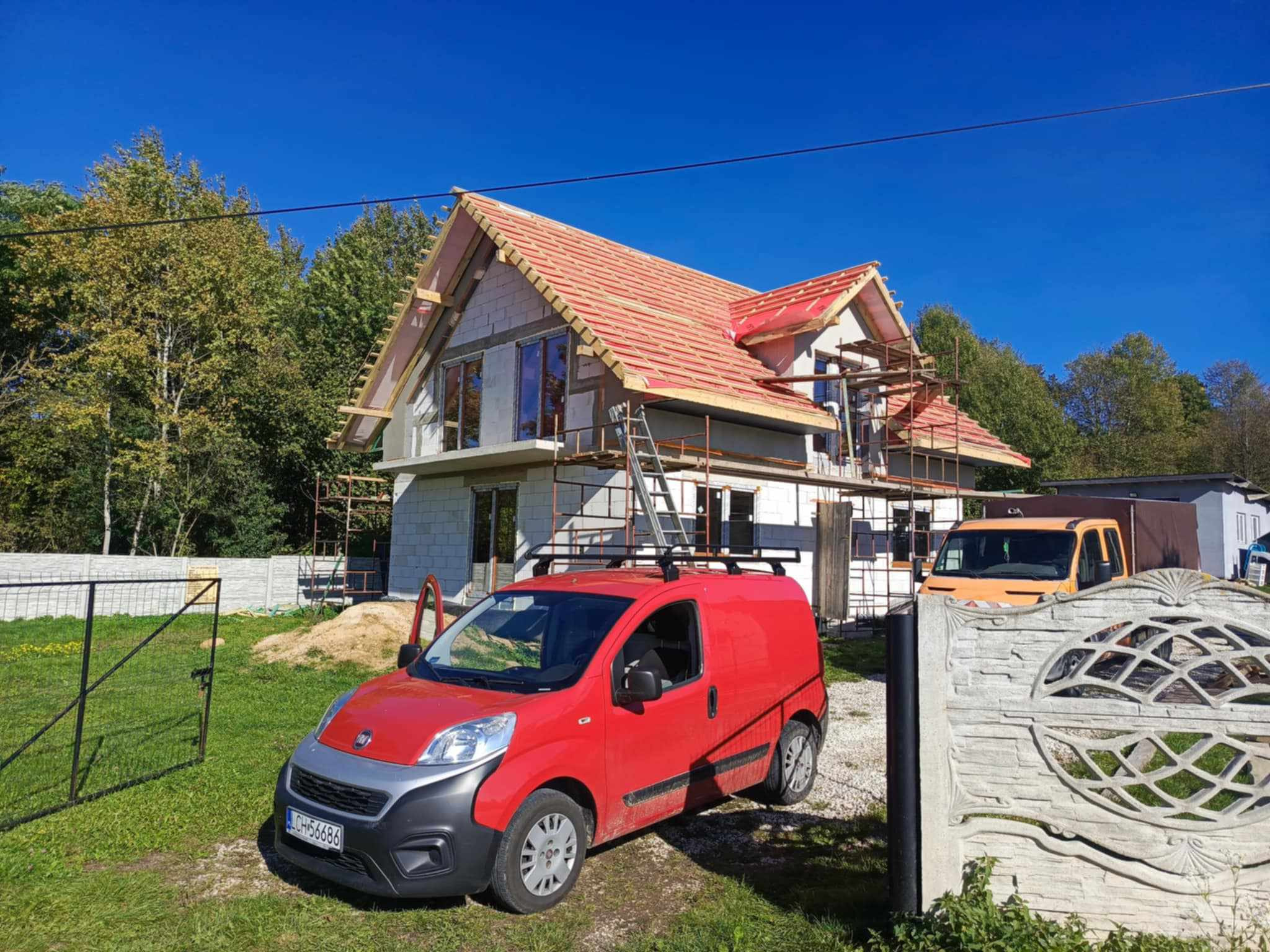 Budowa domu jednorodzinnego w trakcie: widoczna konstrukcja dachu z deskowaniem, częściowo pokryta czerwoną membraną dachową, rusztowania, czerwony samochód dostawczy na pierwszym planie, błękitne...