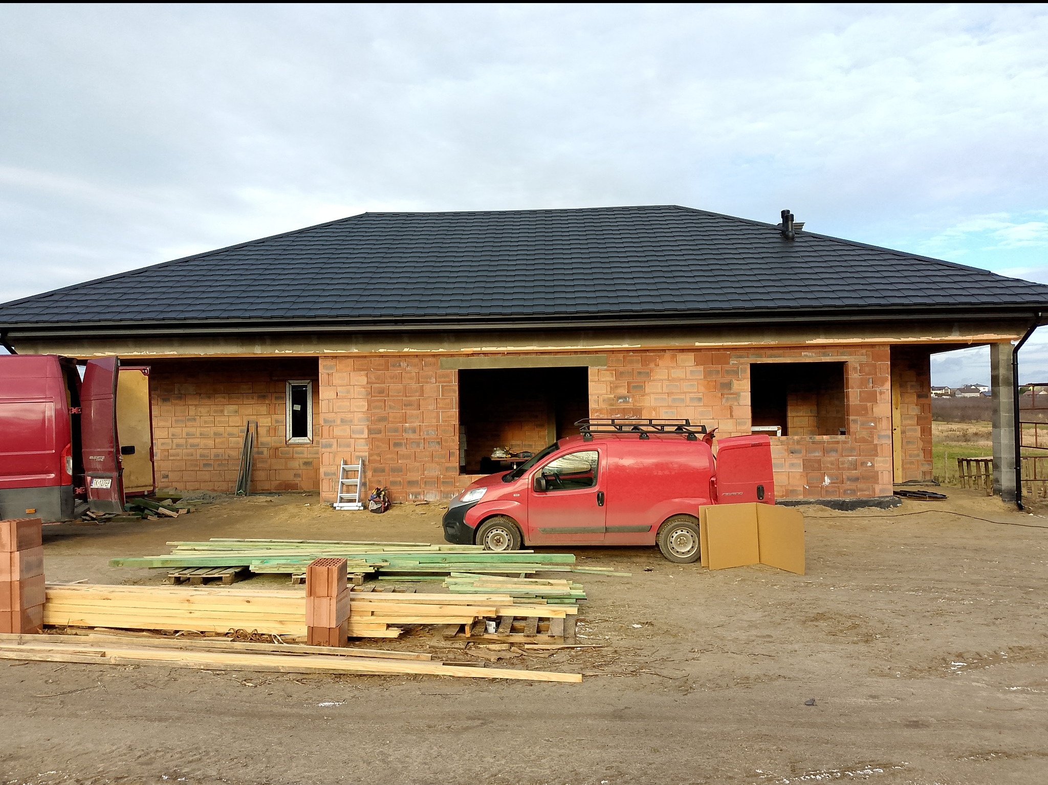 Nowoczesny dom jednorodzinny w trakcie budowy z ciemnym, gotowym pokryciem dachu, czerwonymi samochodami dostawczymi i stertą drewna na nieutwardzonym placu budowy.