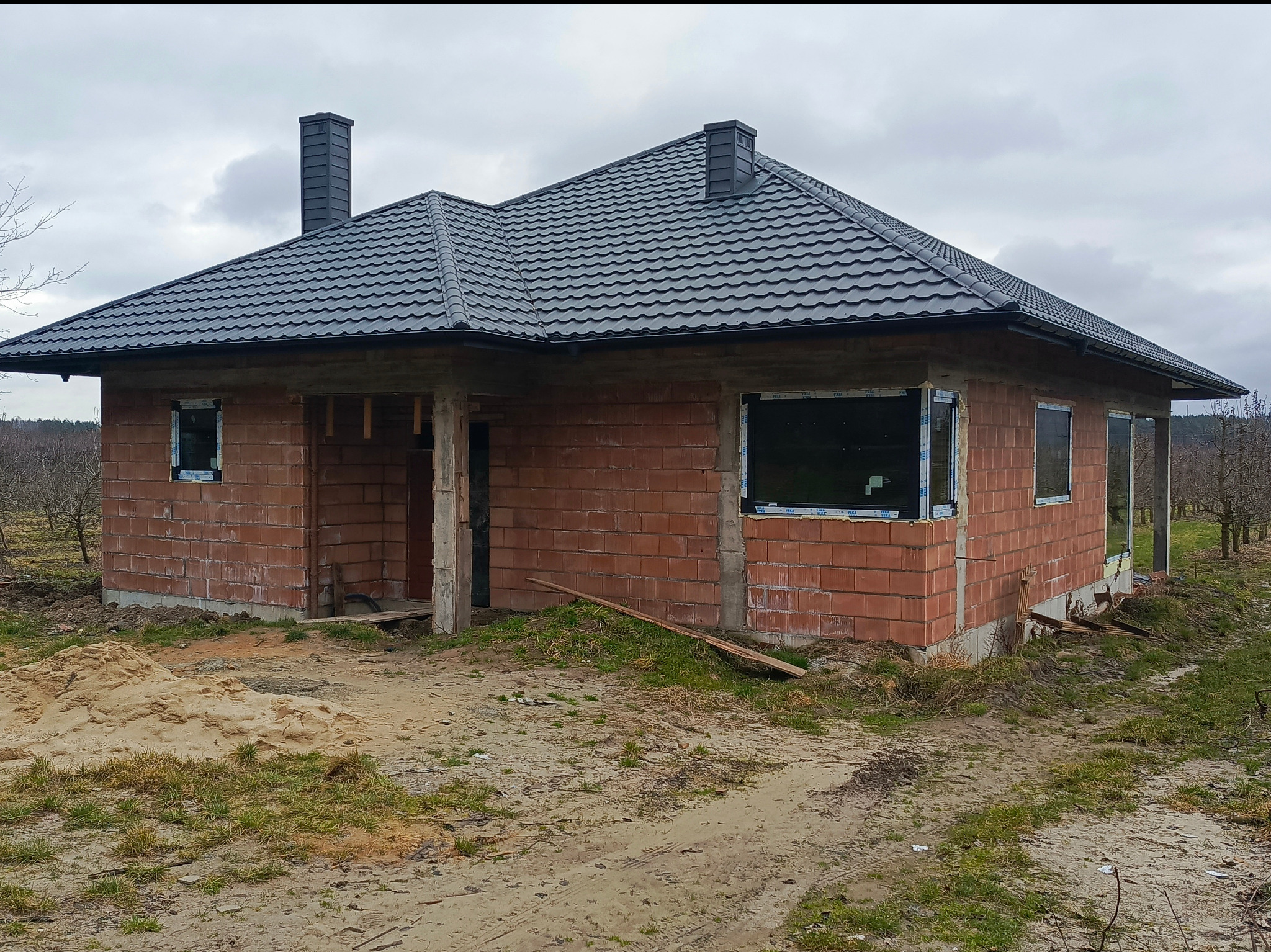 Niedokończony dom jednorodzinny z ciemnoszarą blachodachówką i kominem, widoczne okna z PCV i czerwona cegła, teren wokół domu częściowo zagospodarowany, pochmurne niebo.