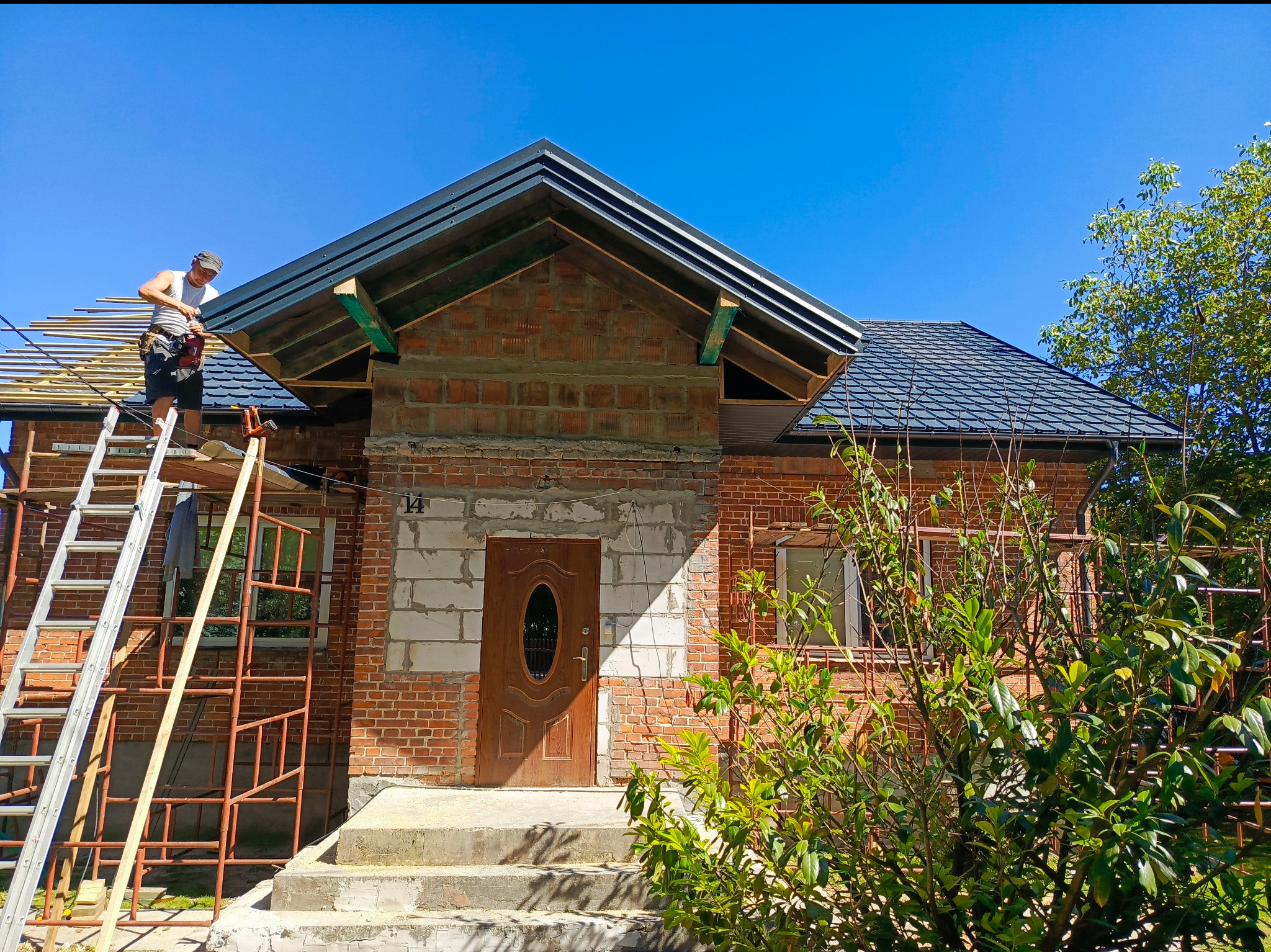 Pracownik na drabinie montuje elementy dachu na nowym domu z czerwonej cegły w słoneczny dzień.