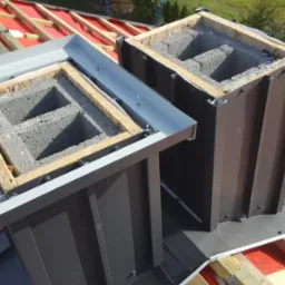 Dwa kominy w trakcie budowy na dachu pokrytym membraną, z widocznymi bloczkami betonowymi i obróbkami blacharskimi w kolorze szarym i brązowym, na tle zieleni.