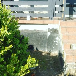 RakBud ogólne pracę remontowo budowlane. - Malowanie elewacji domu w trakcie: pomalowane szare balustrady, widoczna puszka z farbą, fragmenty starej, łuszczącej się farby na cokole. Częściowo odnowione schody.