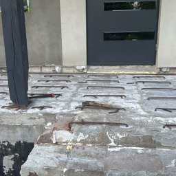 RakBud ogolno pracę remontowo budowlane. - Przygotowanie podłoża pod wylewkę: zbrojenie z prętów, narzędzia (wiertarka, młotek) i czarna kolumna. Widoczne ślady wcześniejszych prac i fragment drzwi.